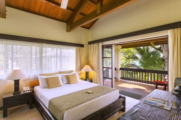 Habitación de hotel con una cama grande, dos mesitas de noche con lámparas, una pequeña mesa con un periódico y un balcón con vistas a árboles.