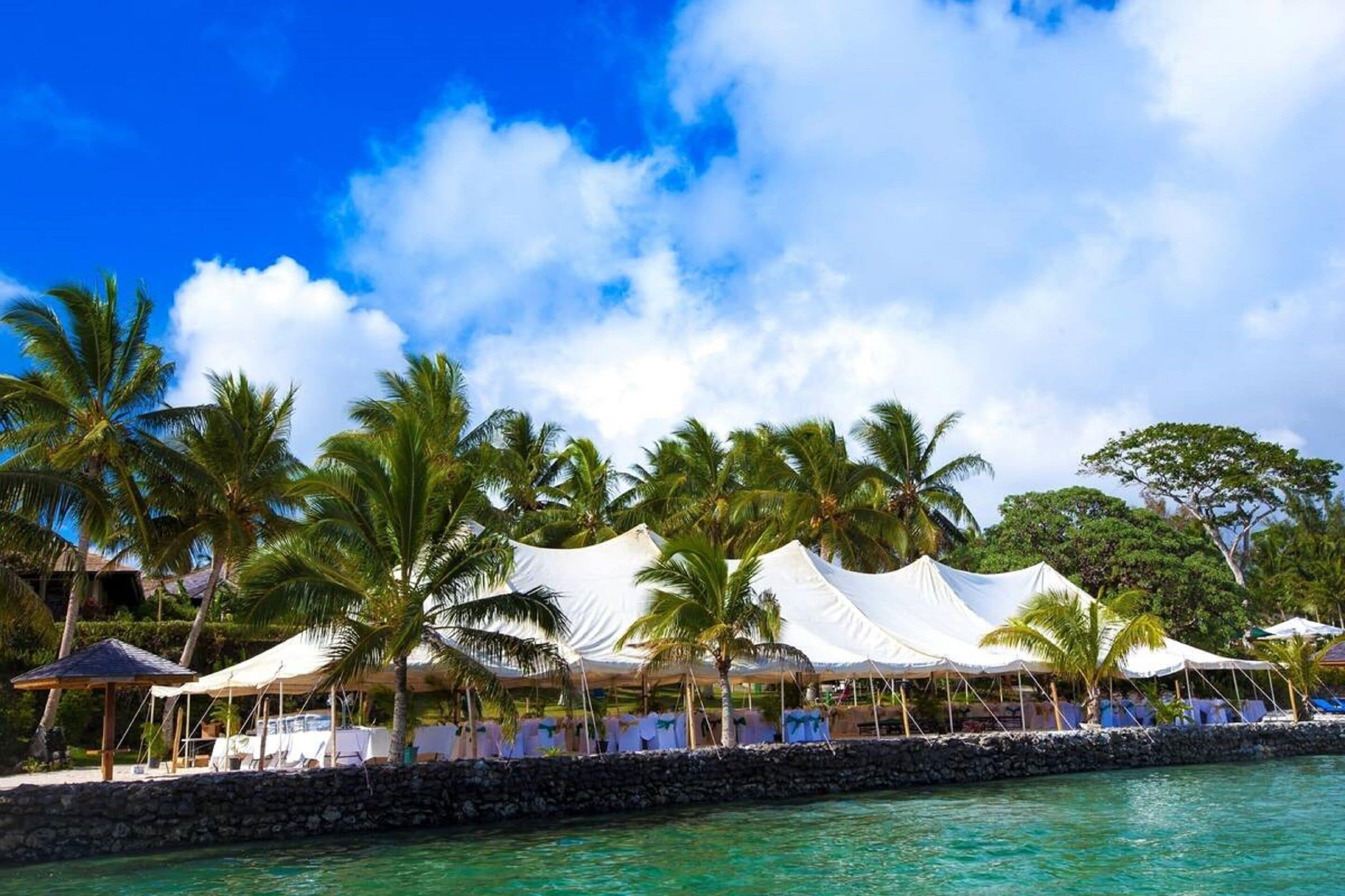 A beachfront resort with white tents, palm trees, and a stone wall.