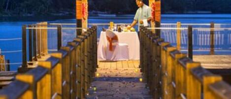 Un muelle de madera que lleva a un pabellón con una mesa dispuesta para dos, con vista a un cuerpo de agua.