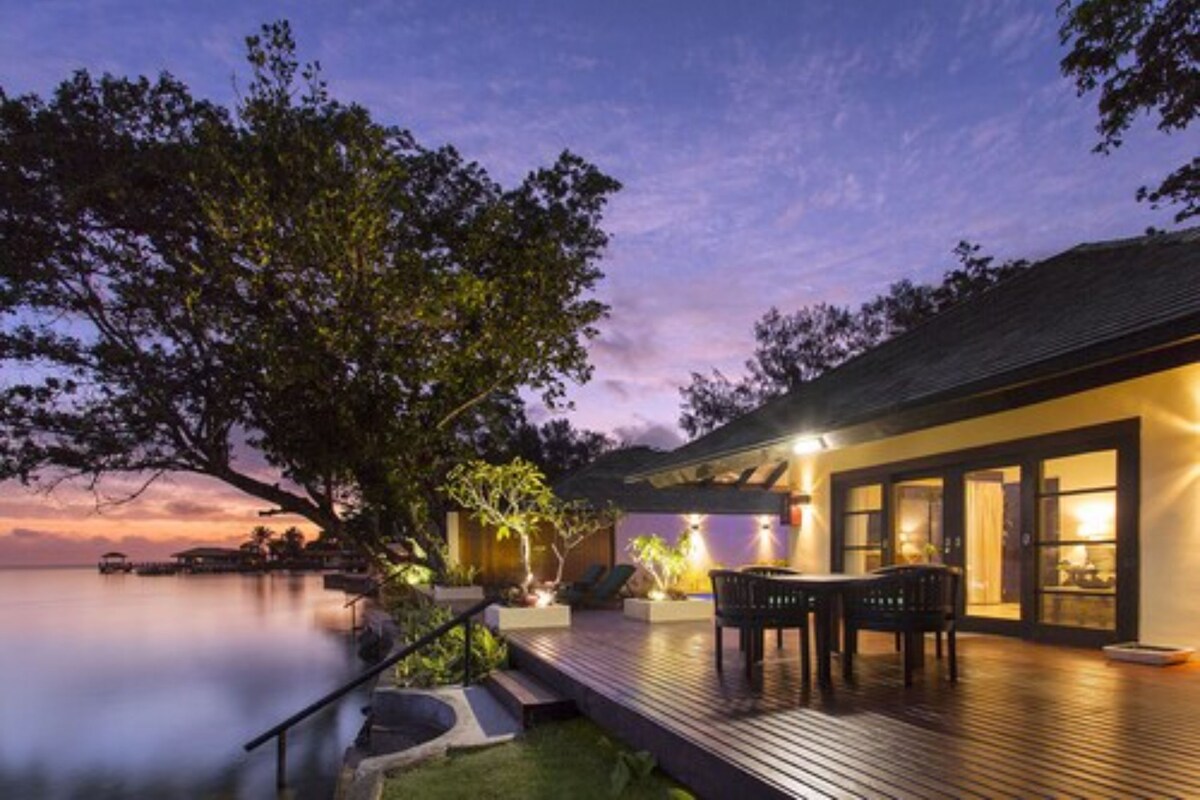 Een pand aan het water bij zonsondergang met een houten terras, buitenmeubels en uitzicht op het water.