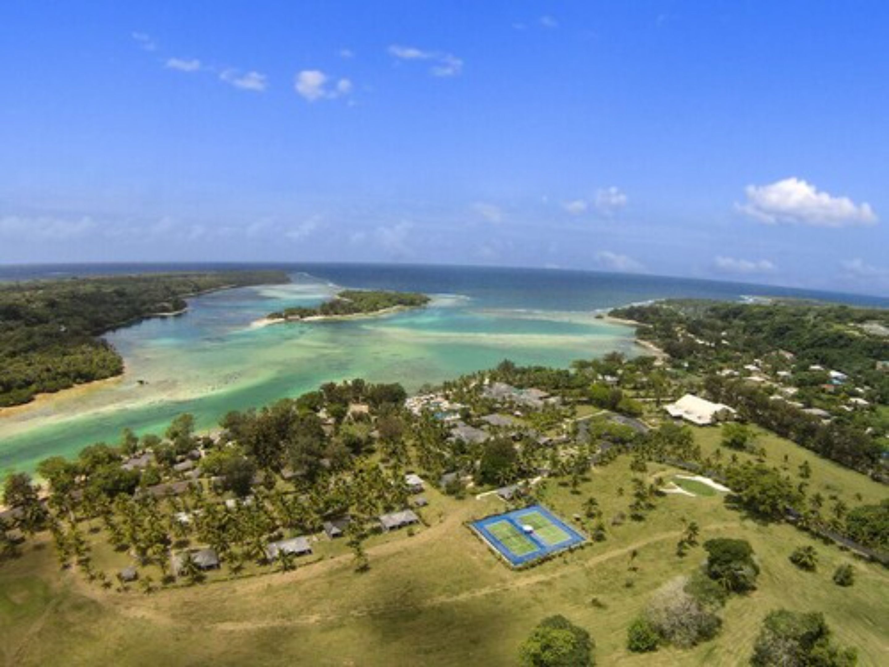 An aerial view of a coastal area with a clear blue sky, a large body of water, and a small settlement with houses and a tennis court.