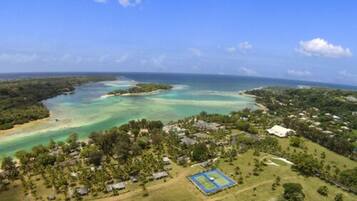An aerial view of a coastal area with a clear blue sky, a large body of water, and a small settlement with houses and a tennis court.