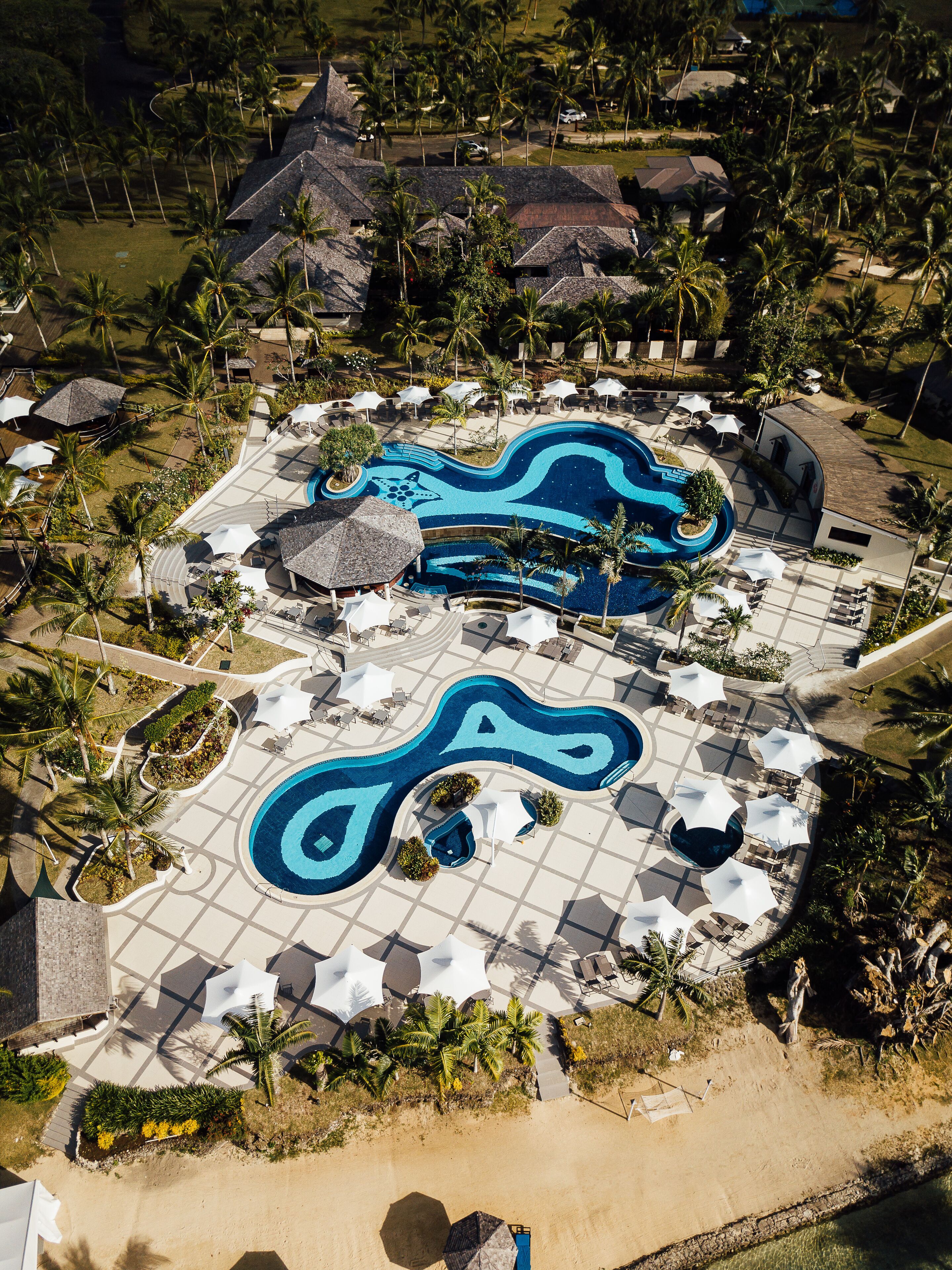 An aerial view of a resort with a large swimming pool, lounge areas, and palm trees.