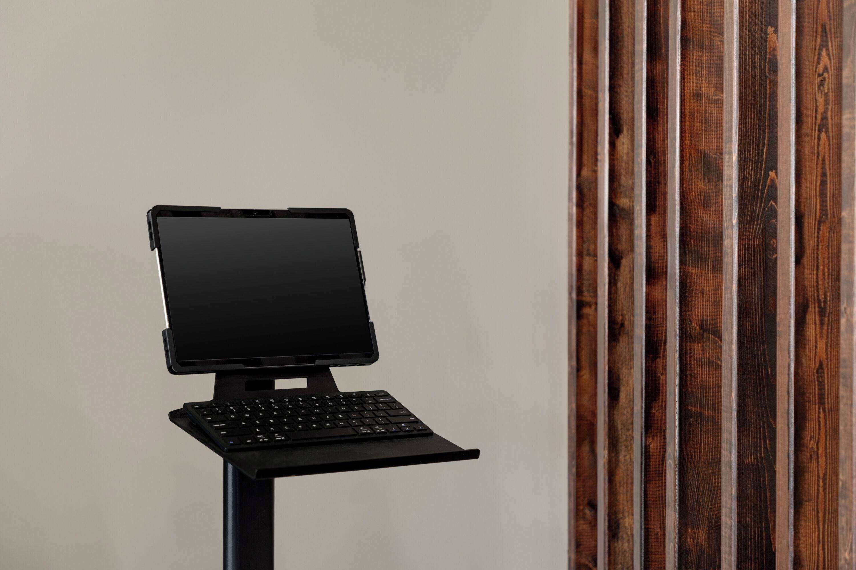 A laptop on a stand with a keyboard, positioned against a plain wall with vertical wooden slats.