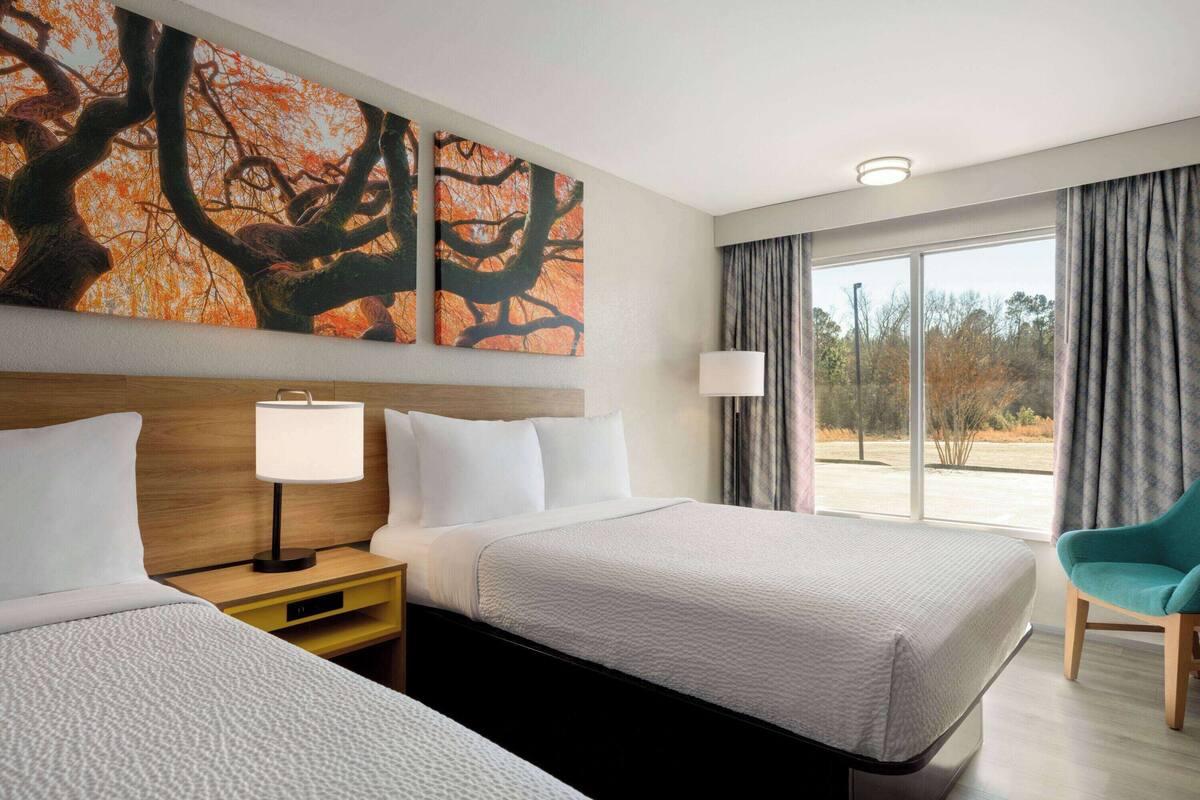 Habitación de hotel con dos camas, un ventanal con cortinas, un cuadro de árboles y una silla color turquesa.