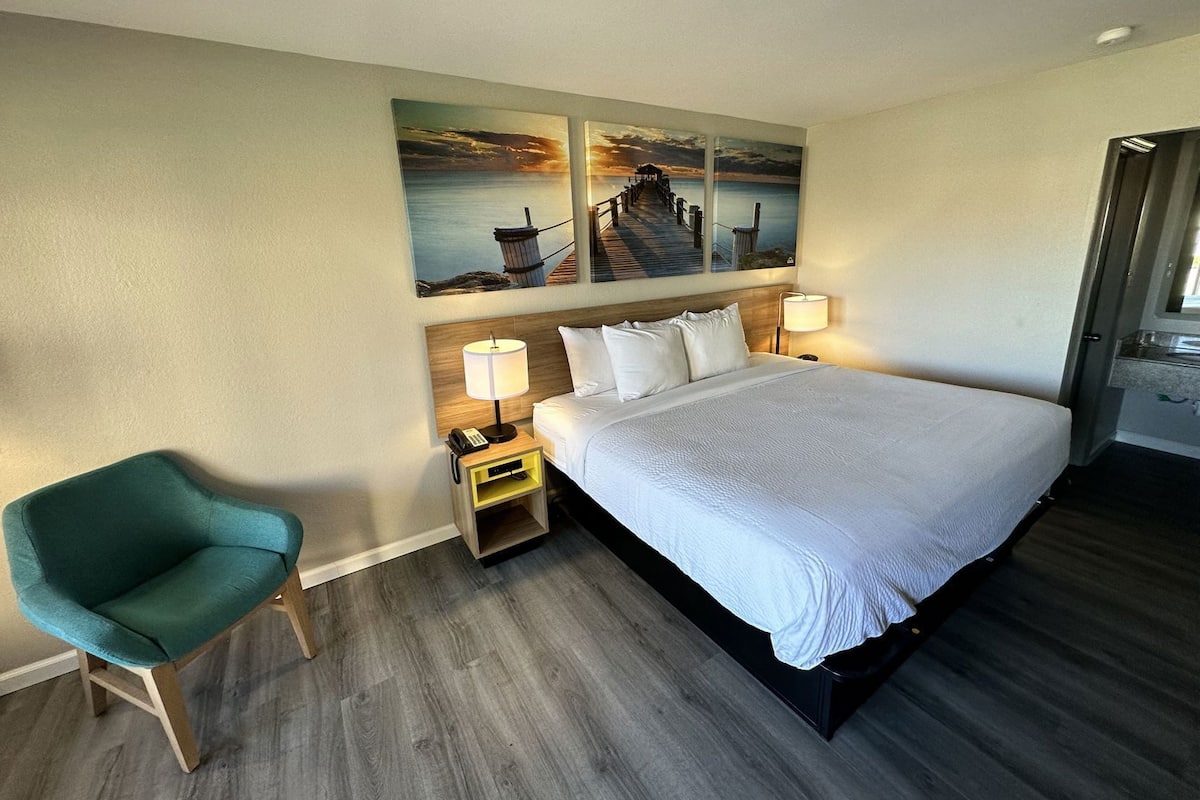 Habitación de hotel con cama, dos lámparas de noche, una mesita de noche, una silla color turquesa y un cuadro de un muelle en la pared.