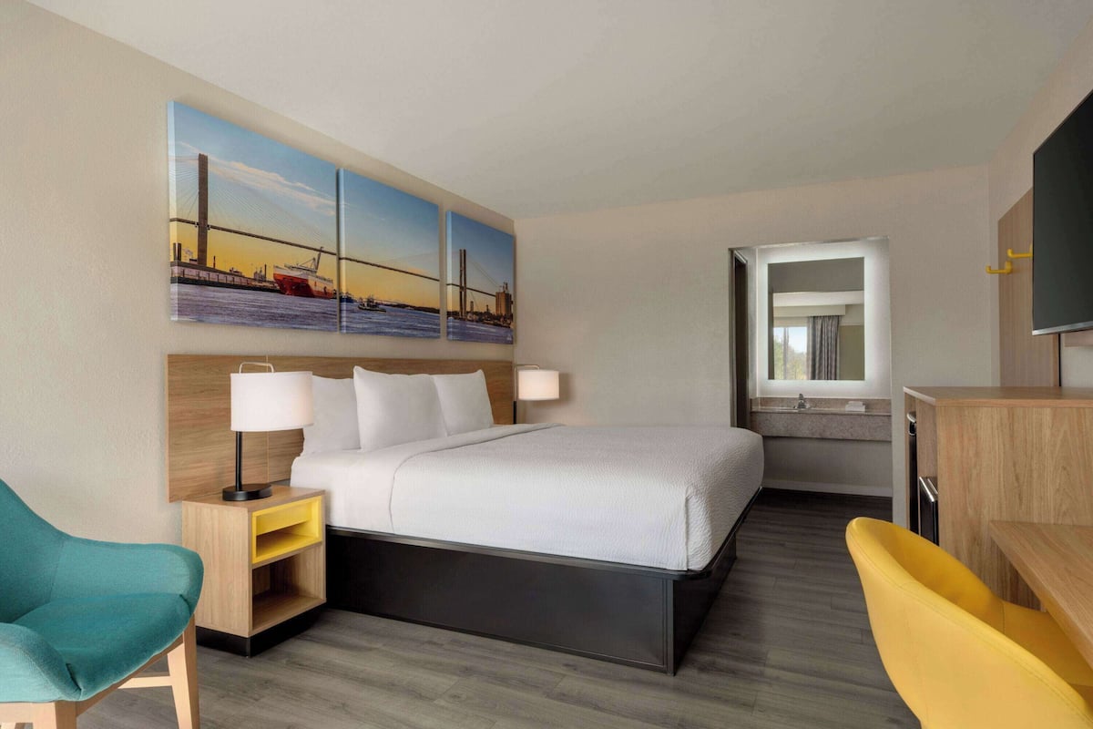 Una habitación de hotel con una cama, un escritorio, una silla, una mesita de noche, una lámpara y un cuadro de un puente.