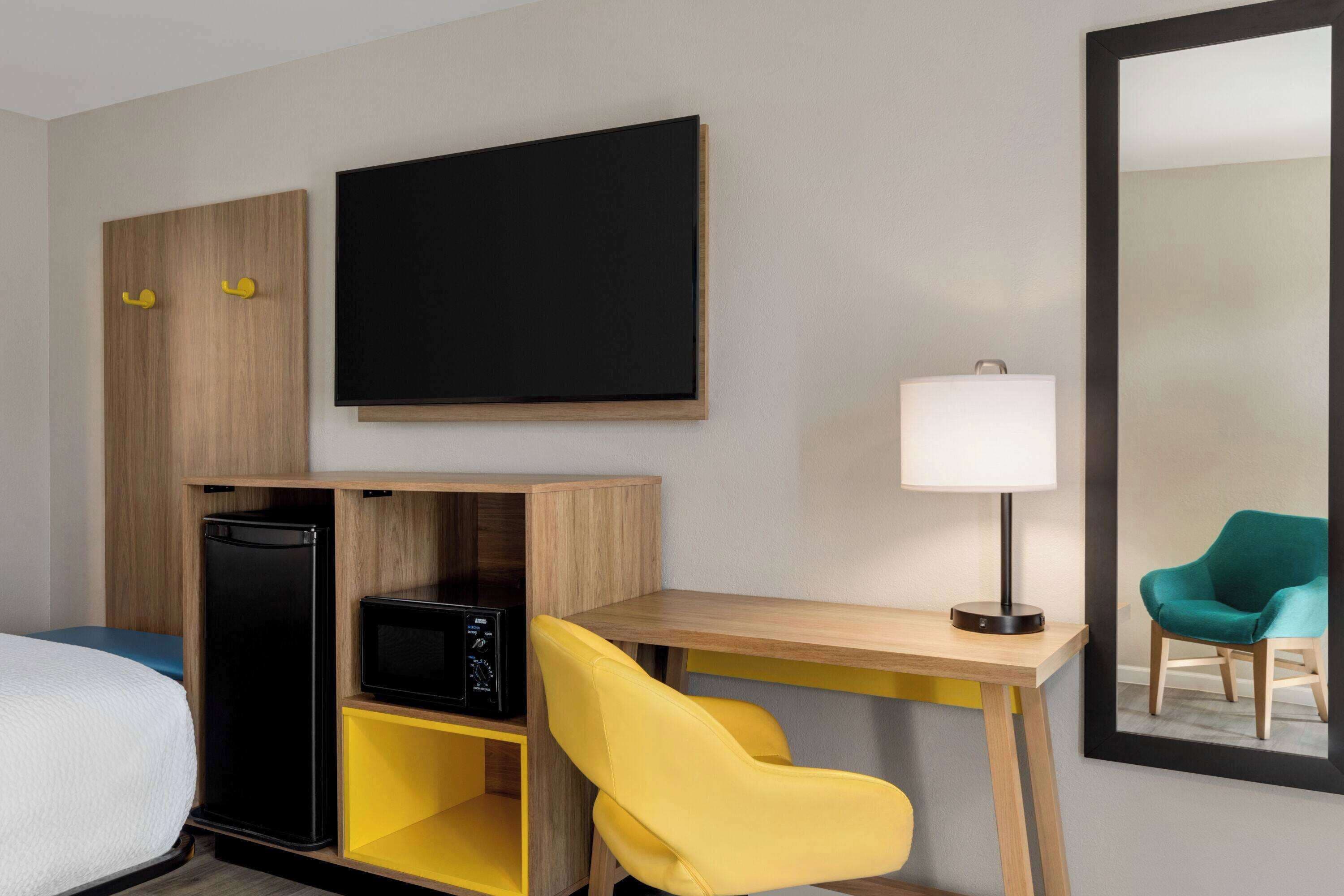 A hotel room with a bed, a desk with a lamp, a TV, a small fridge, a yellow chair, and a mirror reflecting a teal chair.