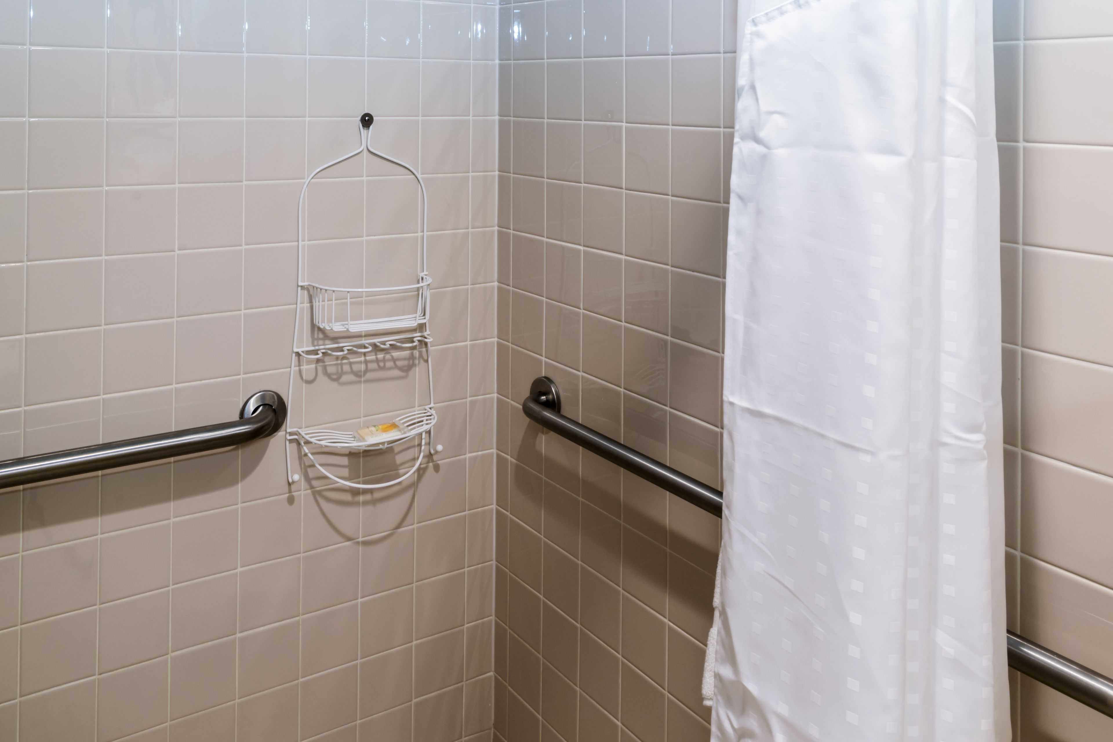 Une salle de bain avec des murs carrelés de couleur beige, un rideau de douche blanc et une barre d’appui.
