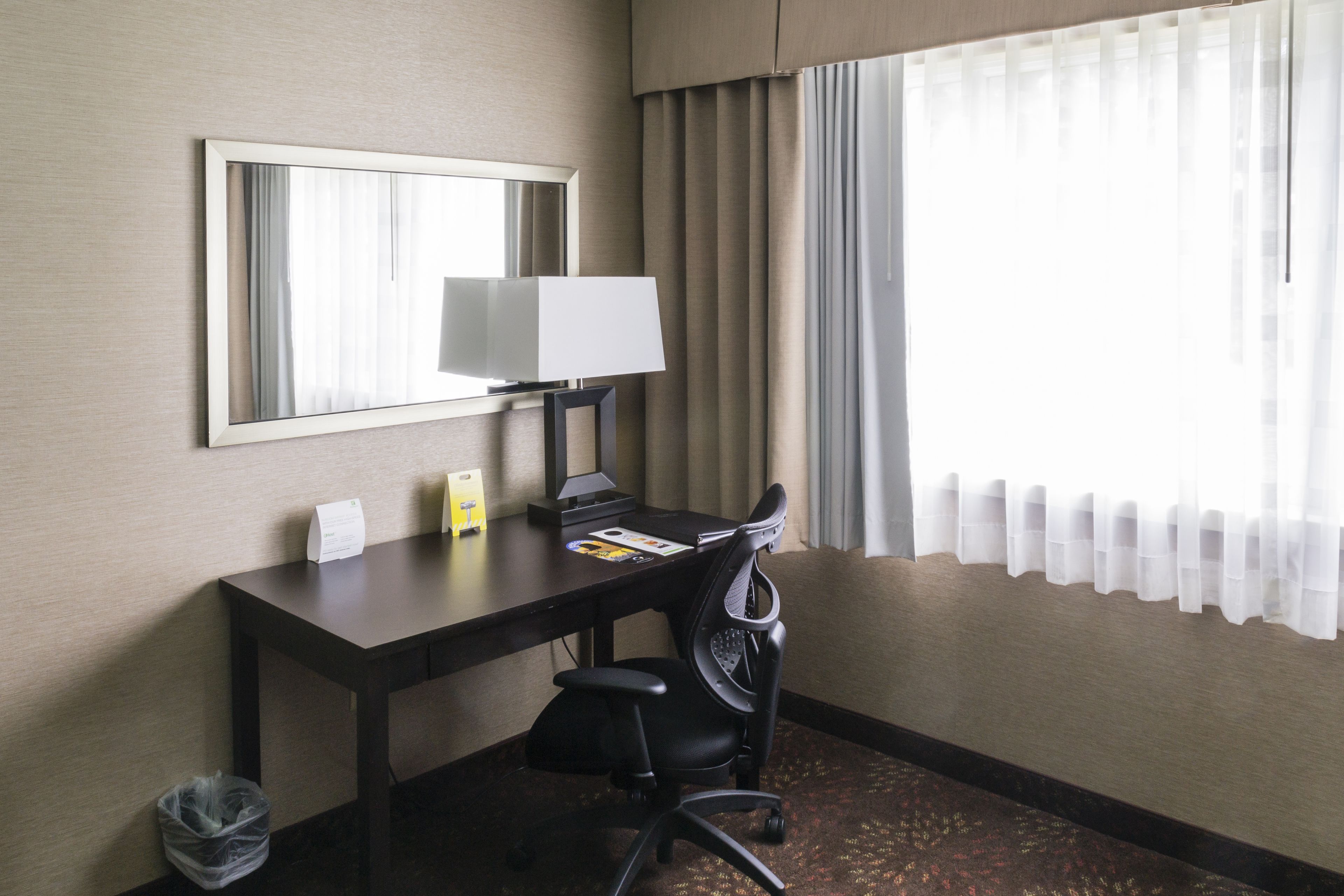 A hotel room with a desk, chair, lamp, and a large mirror on the wall.