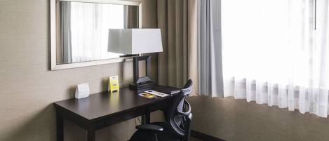 A hotel room with a desk, chair, lamp, and a large mirror on the wall.