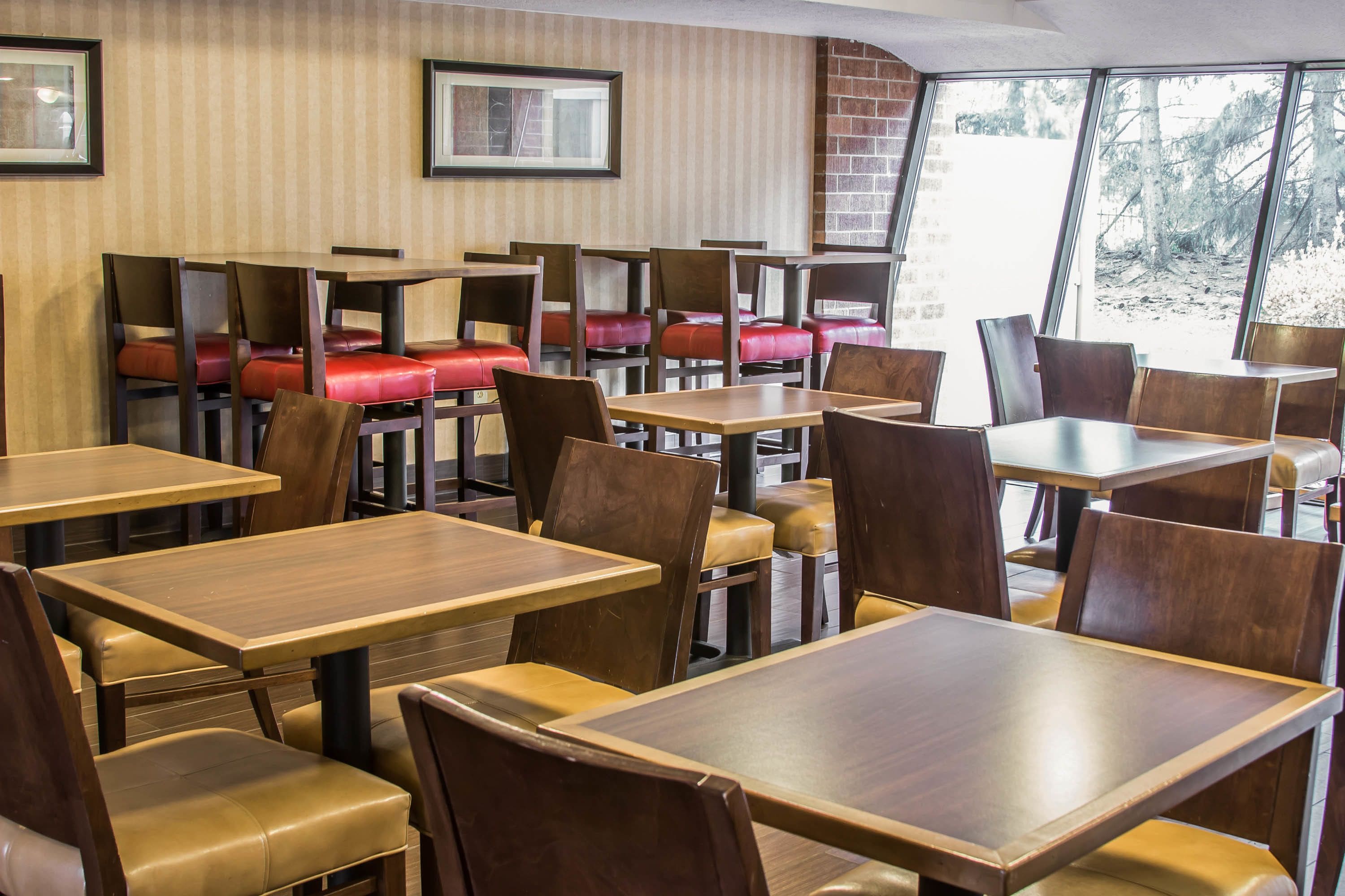 Ein Restaurant mit Holztischen und -stühlen, einige mit roten Kissen. An den Wänden hängen Bilder und es gibt eine Backsteinwand.
