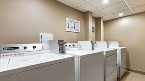 A laundry facility with multiple washing machines and dryers, a control panel, and a bulletin board on the wall. - Comfort Inn Lansing West (Lansing)