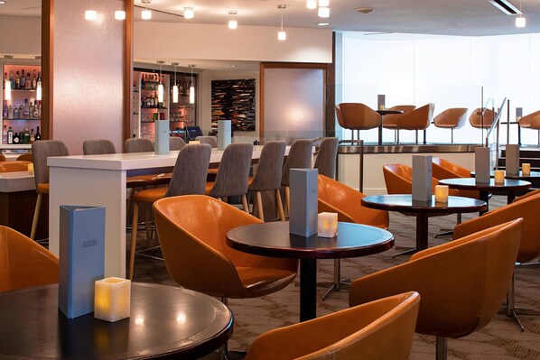 Un restaurante moderno con mesas redondas y sillas naranjas, una zona de bar con estantes llenos de botellas y un amplio ventanal que proporciona luz natural.