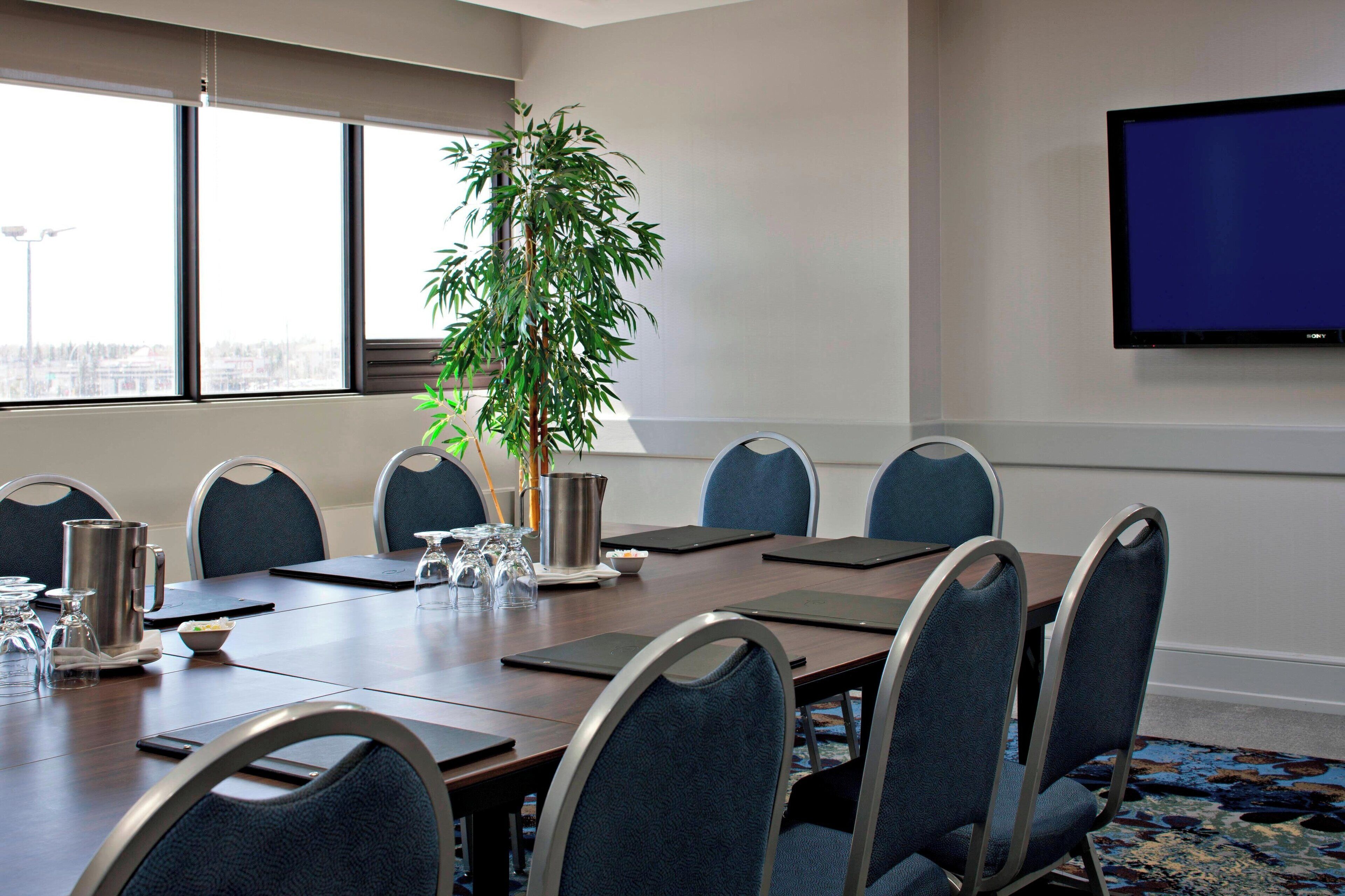 Un salón de conferencias con una gran mesa, sillas, una planta, una cafetera y un televisor en la pared.