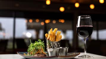 Ein Teller mit Essen, dazu Pommes, Grünzeug, ein Glas Rotwein und eine kleine Metalltasse auf einem Tisch in einem schwach beleuchteten Raum mit Pendelleuchten.