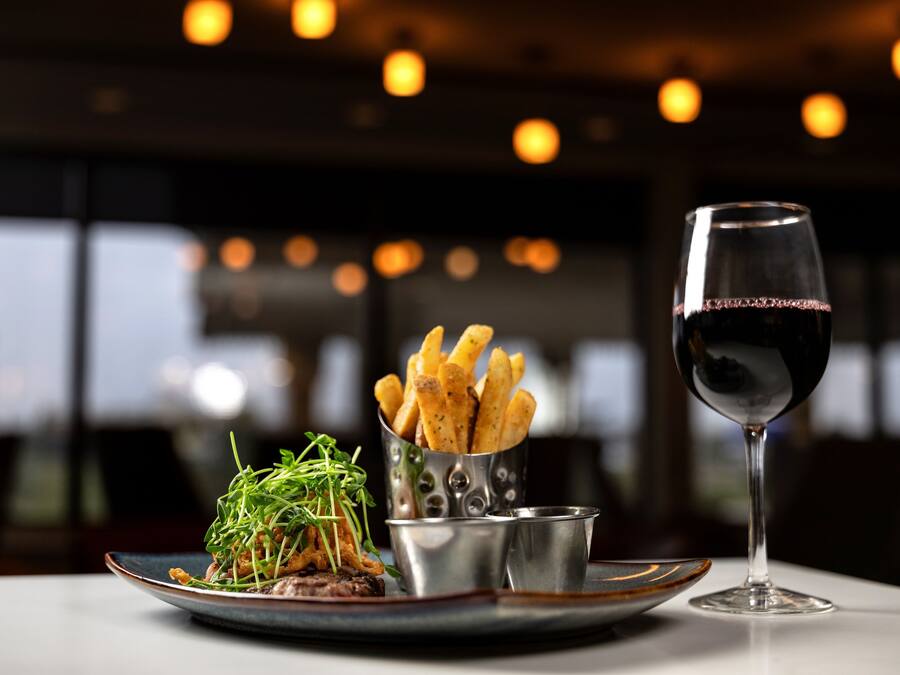 Ein Teller mit Essen, dazu Pommes, Grünzeug, ein Glas Rotwein und eine kleine Metalltasse auf einem Tisch in einem schwach beleuchteten Raum mit Pendelleuchten.