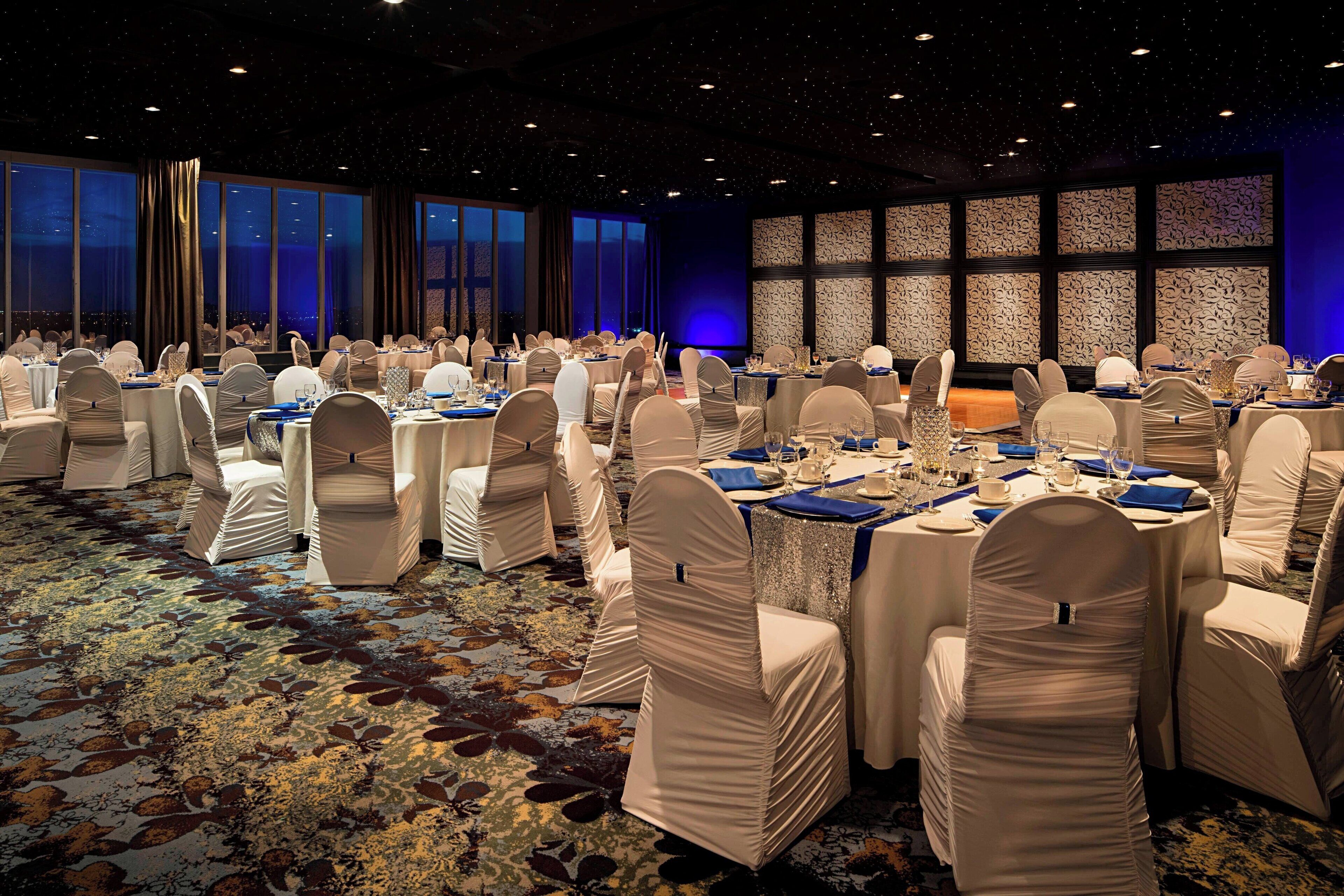 A banquet hall with round tables and chairs, large windows, and a patterned carpet.