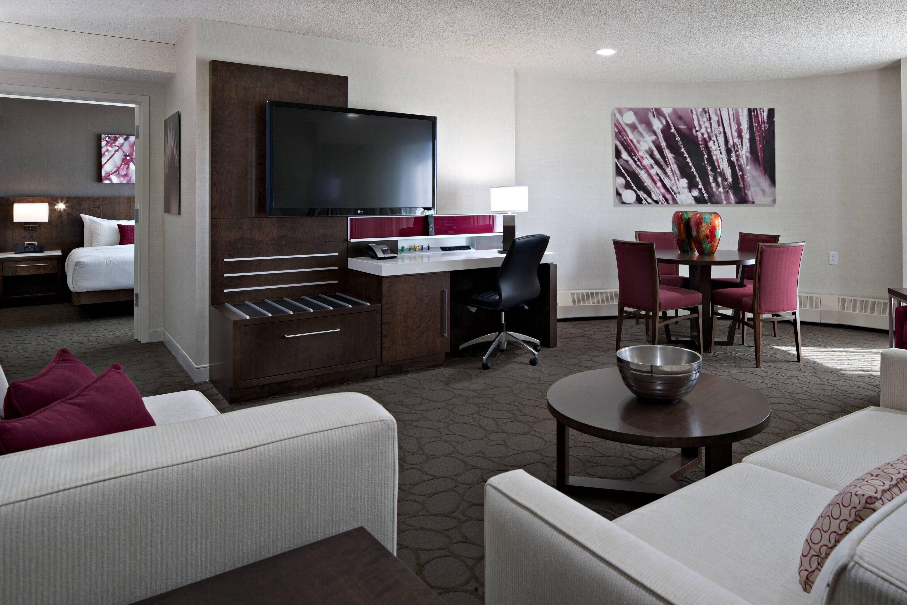 A hotel room with a bed, a desk with a computer, a TV, a dining table with chairs, and a large abstract painting on the wall.