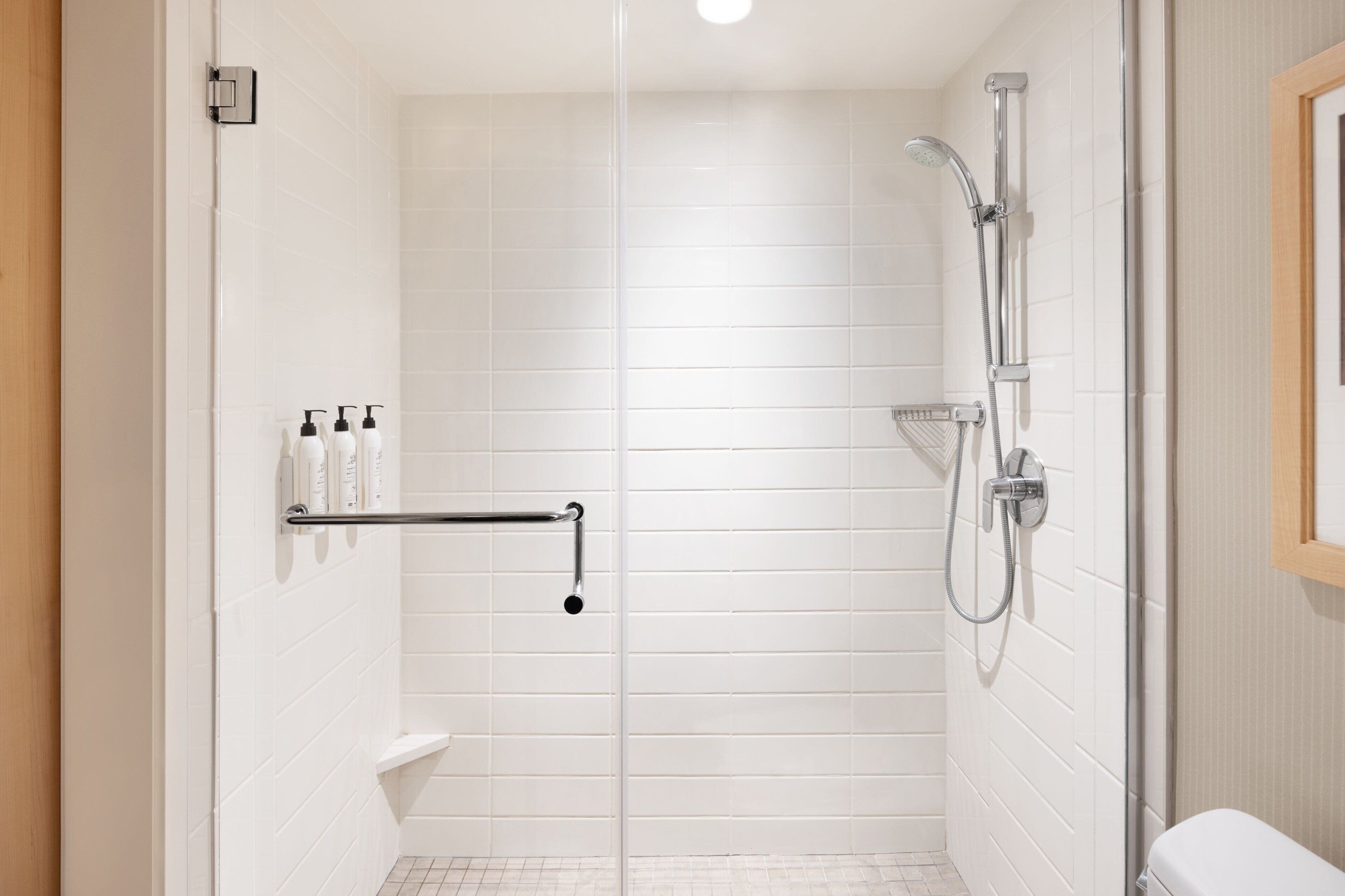 A modern bathroom with white tiled walls, a glass shower enclosure, a toilet, and a towel rack with toiletries.