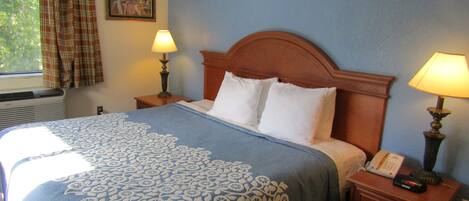 Una cama bien hecha con colcha de motivos florales, dos lámparas de noche, una mesita con teléfono y una ventana con cortinas a cuadros.
