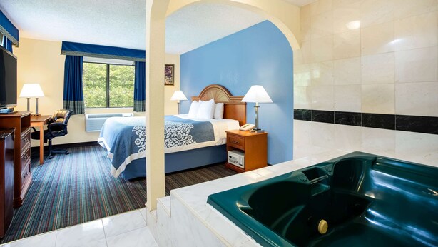 A hotel room with a bed, a desk, a chair, a desk lamp, a nightstand, a telephone, a window with curtains, and a bathtub. - Days Inn by Wyndham Runnemede Philadelphia Area (Runnemede)