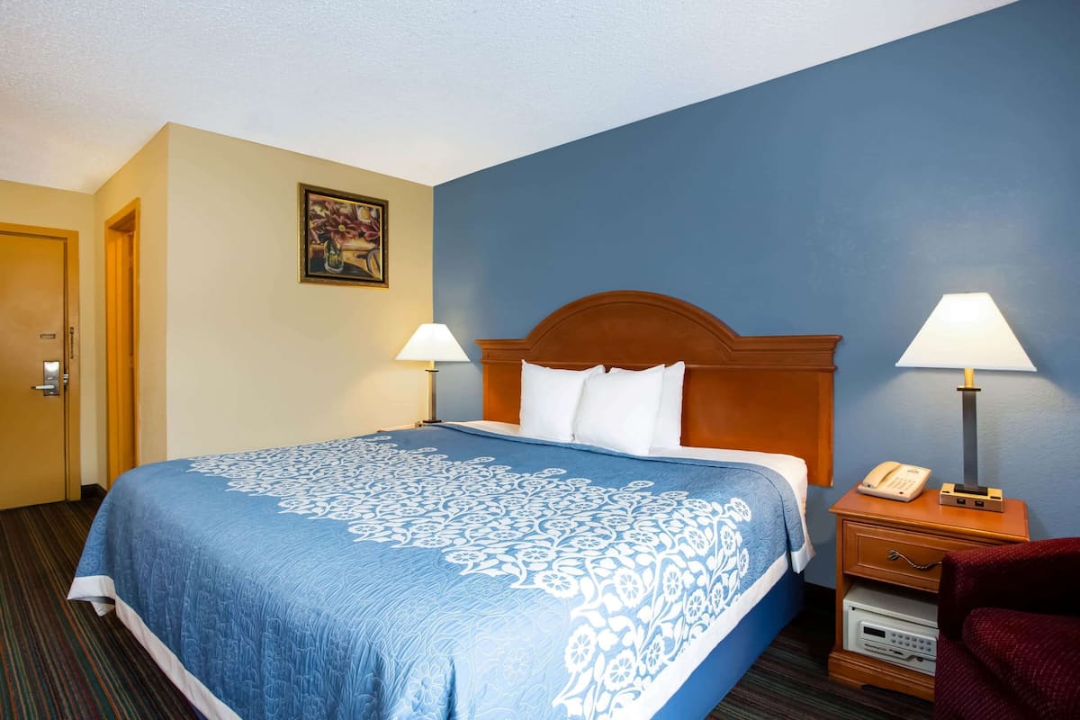 A hotel room with a large bed, two bedside lamps, a wooden headboard, a painting on the wall, and a door to the left.