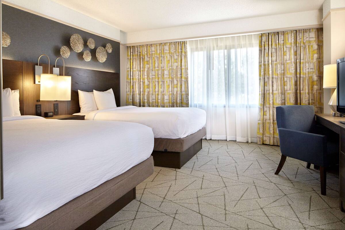 Une chambre d’hôtel avec deux lits, un bureau, une chaise et une grande fenêtre avec des rideaux.