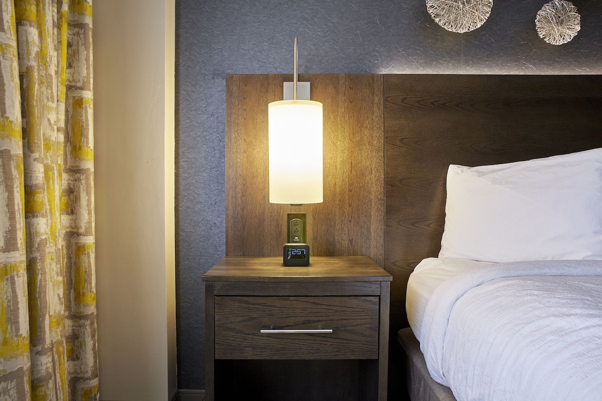 A neatly made bed with white linens, a wooden nightstand with a digital clock, and a wall-mounted lamp.