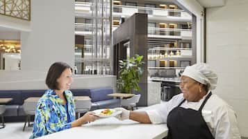 Un moderno vestíbulo de hotel con un área de descanso, una planta grande y un mostrador donde un camarero sirve un plato de comida a un huésped.