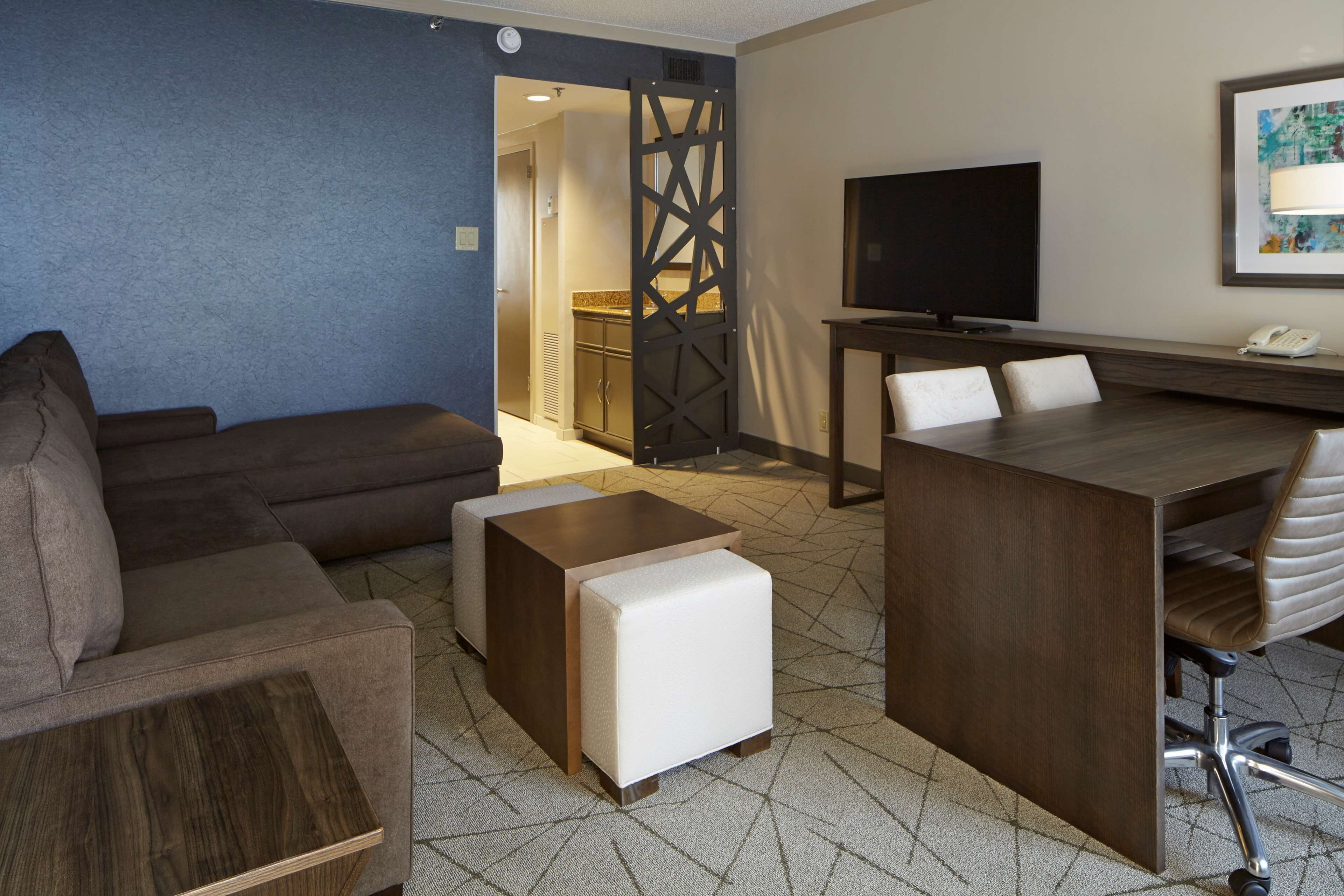 A modern hotel room with a sofa, ottoman, wooden coffee table, and a desk with a chair. A TV and phone are mounted on the wall.