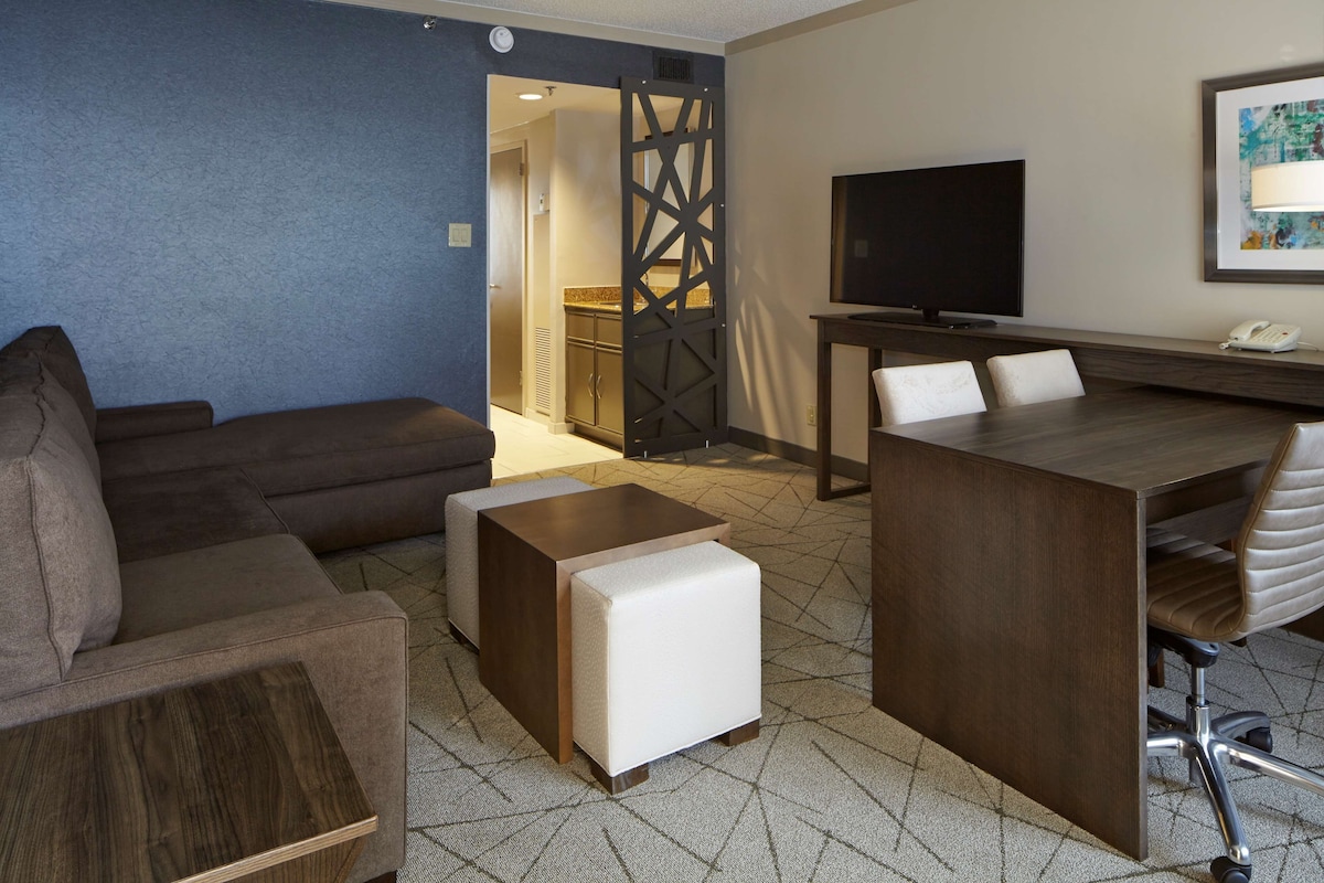 A modern hotel room with a sofa, ottoman, wooden coffee table, and a desk with a chair. A TV and phone are mounted on the wall.
