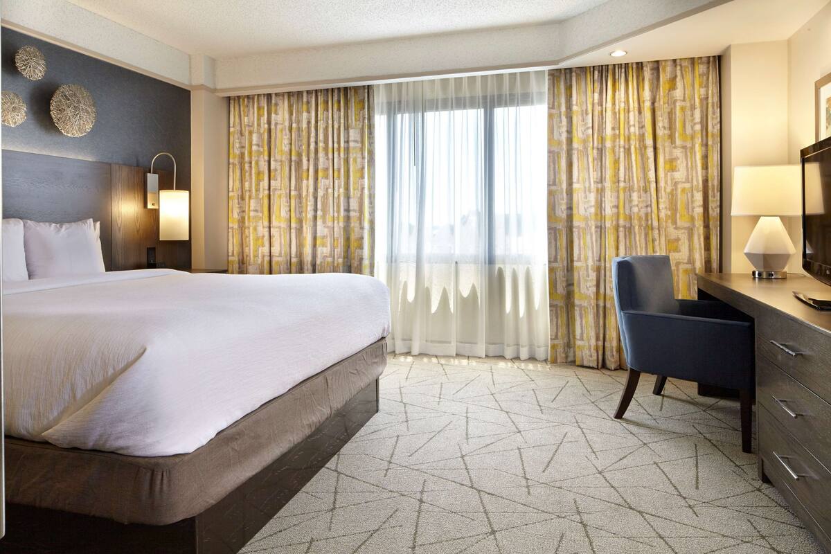 Une chambre d’hôtel avec un grand lit, un bureau avec une chaise, une télévision et une fenêtre avec des rideaux.