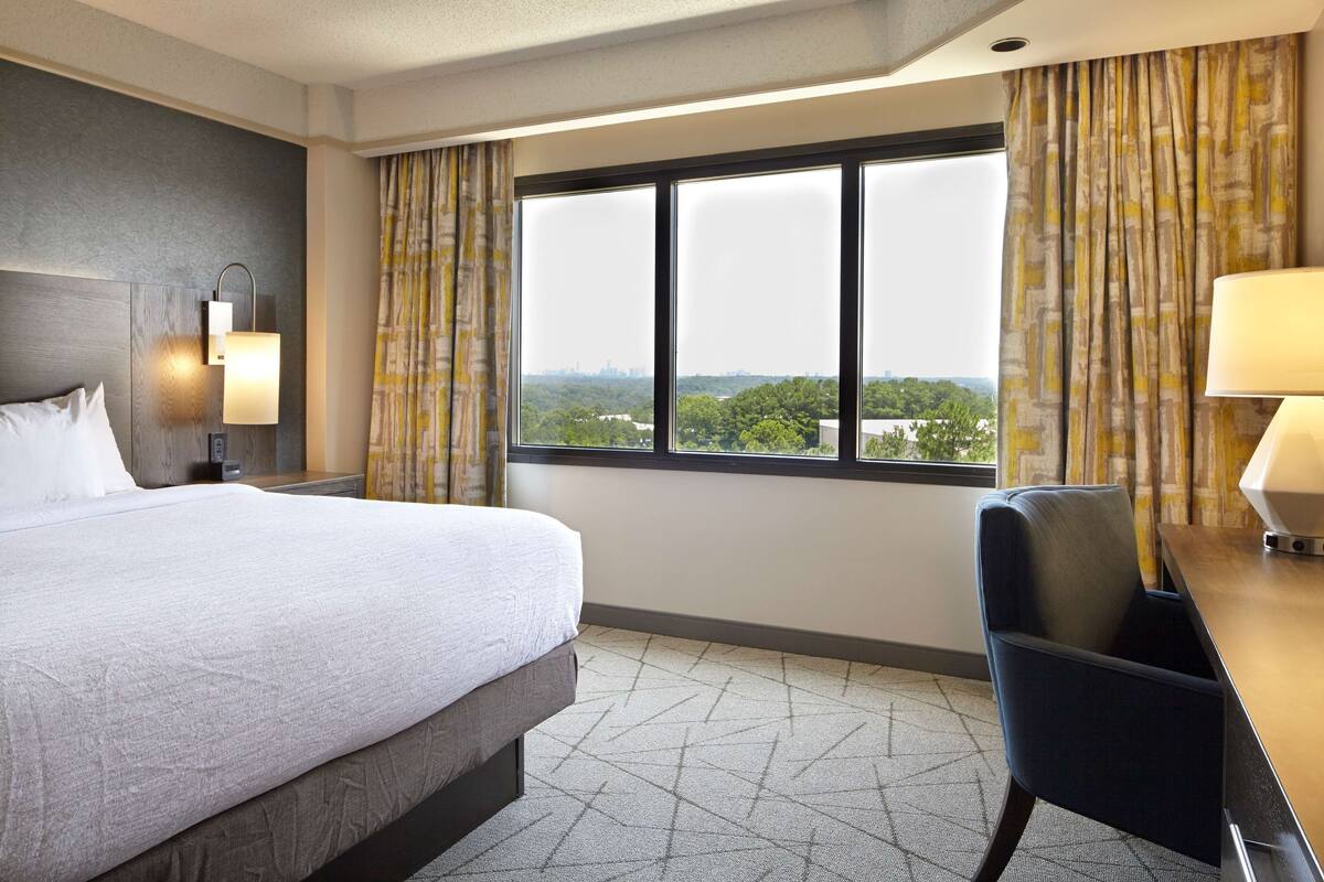 Une chambre d’hôtel avec un grand lit, un bureau, une chaise, une lampe, une table de chevet et une vue sur la ville par la fenêtre.