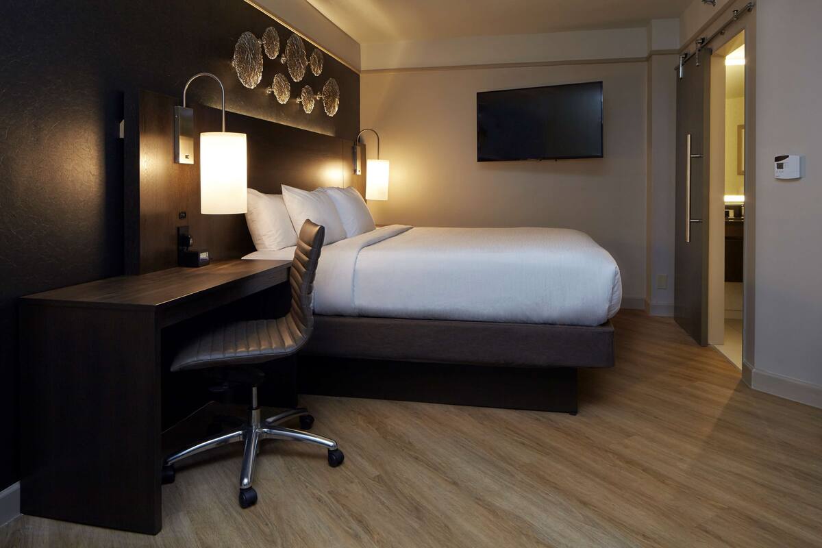 Une chambre d’hôtel moderne équipée d’un lit, d’un bureau, d’une chaise et d’un téléviseur à écran plat fixé au mur.