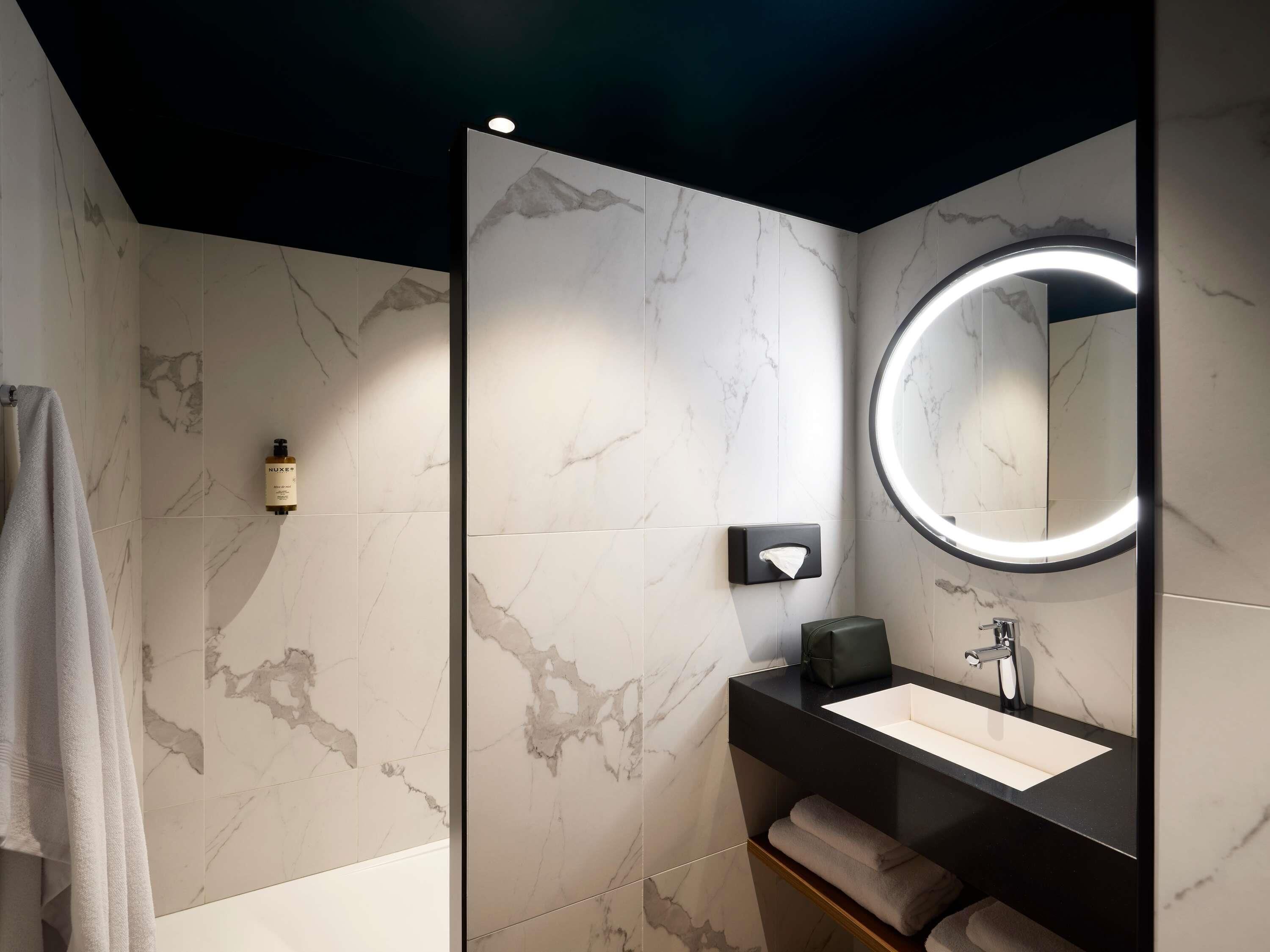 Een moderne badkamer met een marmeren wand, een ronde spiegel met verlichting, een zwarte wastafel en een handdoekhouder.