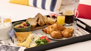 Bandeja de desayuno con croissants, pan, queso y un bol de fruta sobre una cama con almohadas.