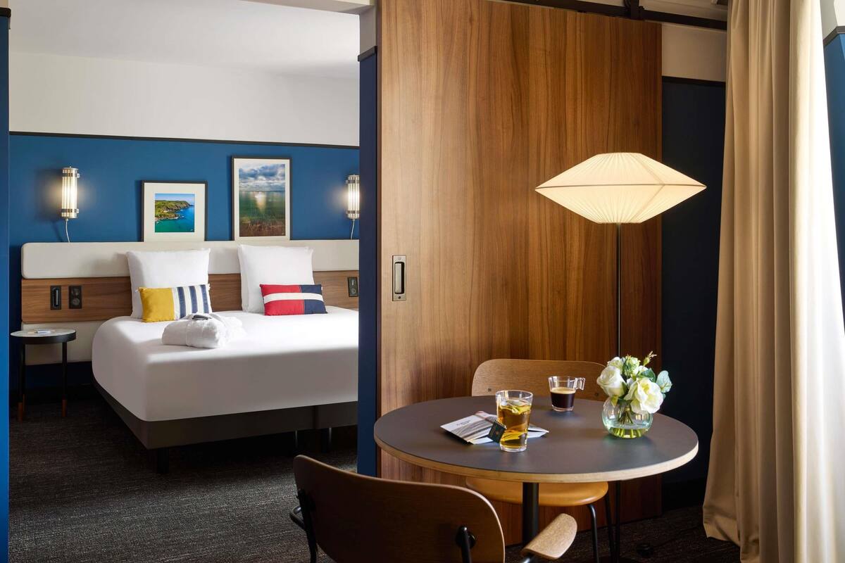 Habitació d'hotel amb llit, taules de nit, una taula rodona amb cadires, una làmpada i un vas amb flors.