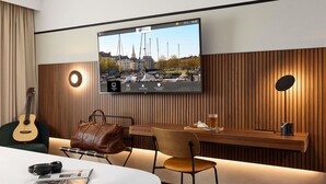 Habitación de hotel con una televisión que muestra un paisaje urbano, una guitarra, un bolso de mano, un escritorio de madera y una silla.