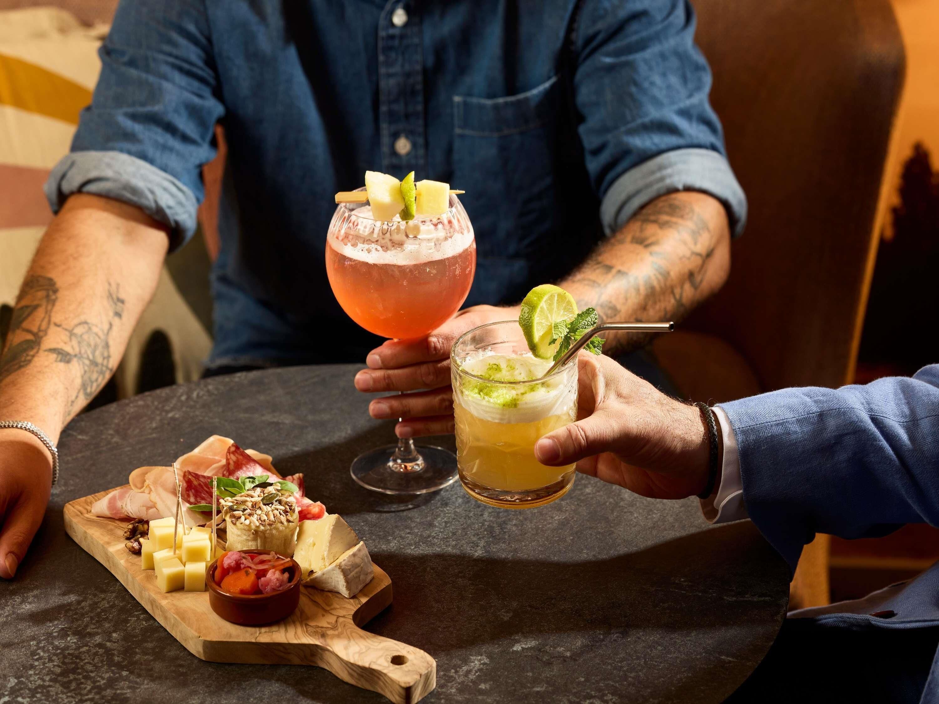 Une table sur laquelle on trouve une planche en bois garnie d’un assortiment de fromages, un verre de cocktail et un verre de boisson fraîche.