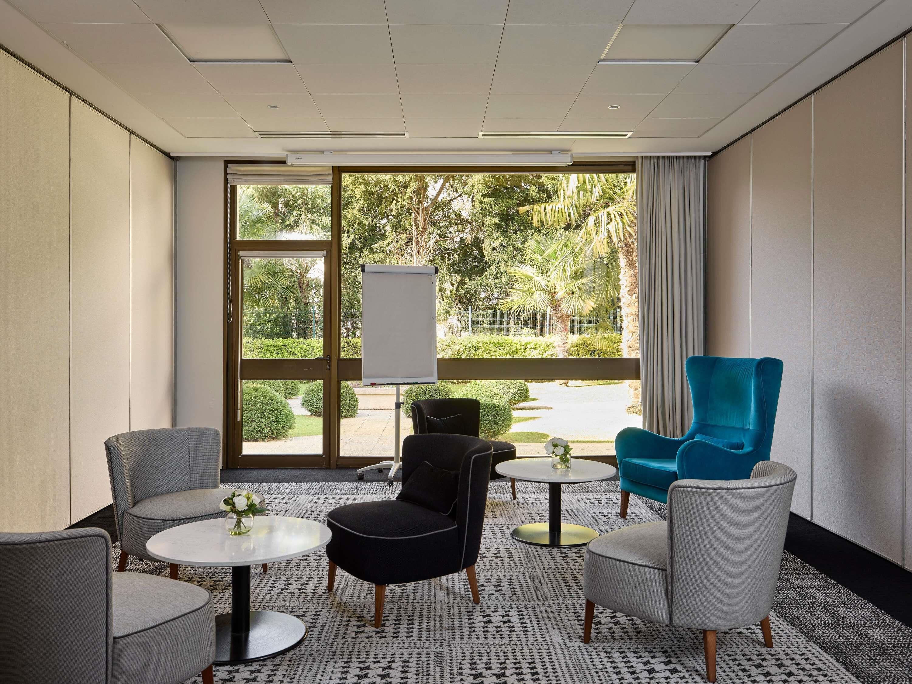 Un salón de conferencias con un amplio ventanal, una pizarra y muebles modernos, incluyendo un sillón azul, sillones grises y un otoman negro.