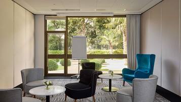 Un salón de conferencias con un amplio ventanal, una pizarra y muebles modernos, incluyendo un sillón azul, sillones grises y un otoman negro.