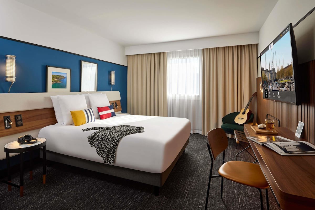 Une chambre d’hôtel avec un lit, un bureau, une chaise, une télévision et une fenêtre avec des rideaux.