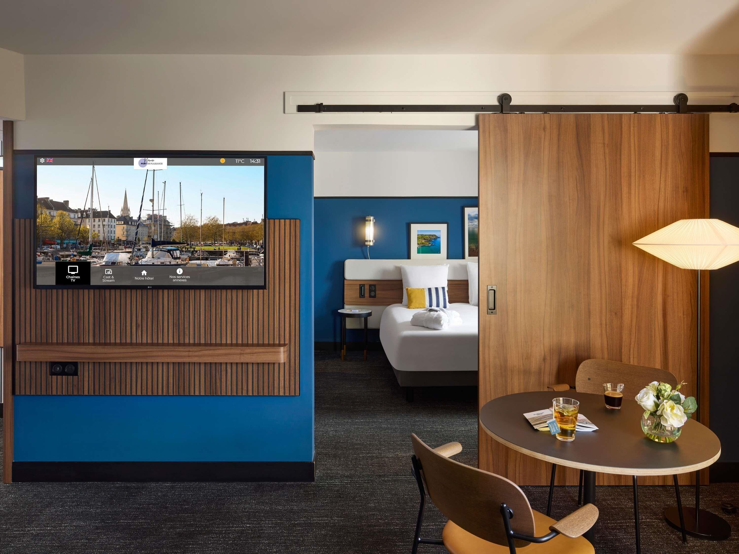 Habitación de hotel con televisión mostrando un paisaje urbano, una cama, un escritorio, una silla y una mesa con un jarrón de flores.