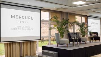 A conference room with a large screen displaying 'MERCURE HOTELS CAEN CENTRE PORT DE PLAISANCE.' There are two chairs, a table, and a staircase leading to a balcony with a view.