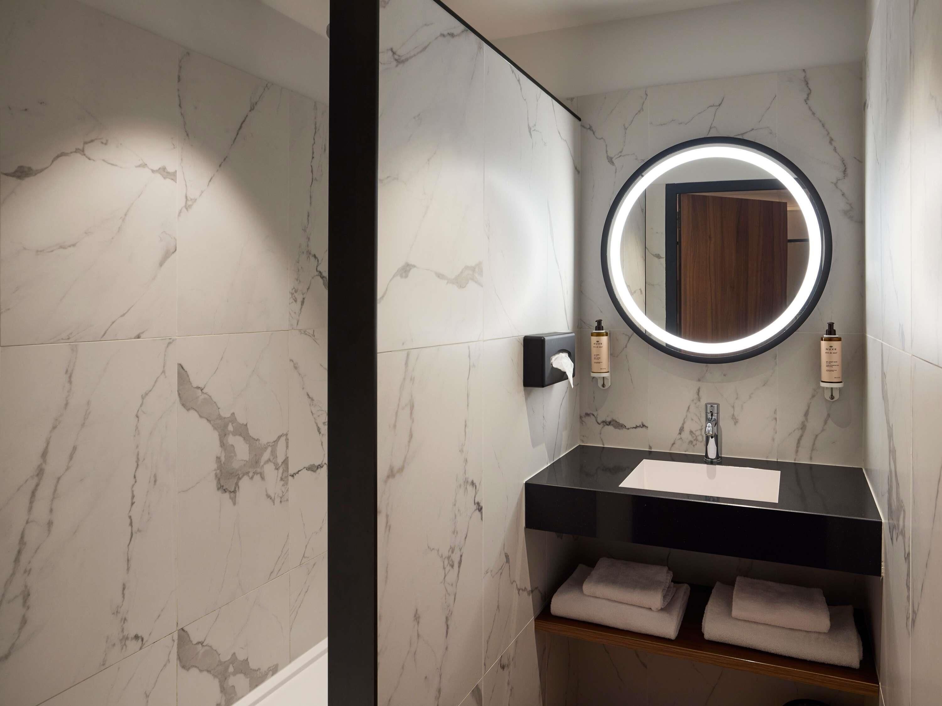 A bathroom with marble walls, a round mirror with a light, a black sink, and two bottles of lotion.