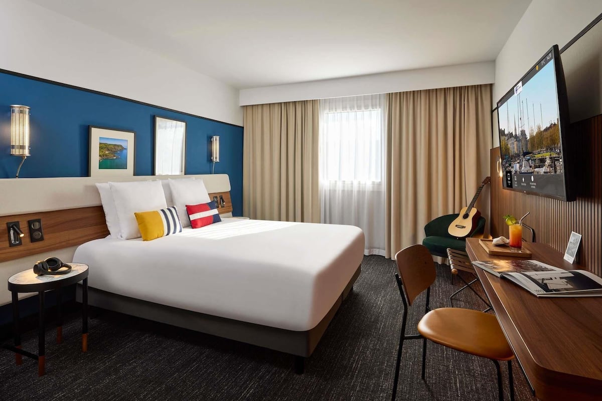 A hotel room with a bed, a desk, a chair, a TV, and a window with curtains.
