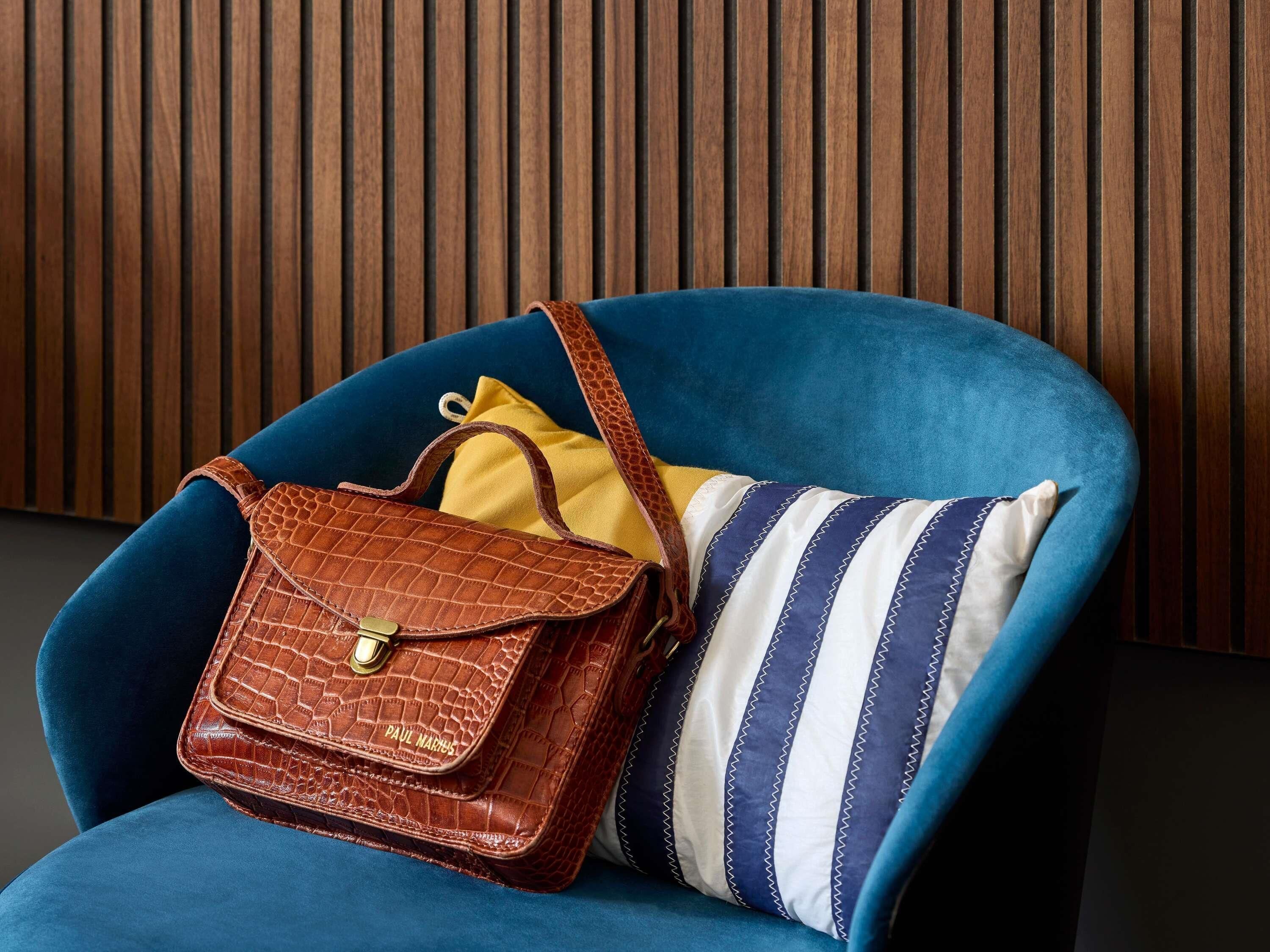 Un fauteuil bleu, accompagné d’un sac à main en cuir marron et d’un coussin à rayures, le tout placé devant un panneau en bois.
