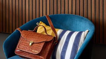 Un fauteuil bleu, accompagné d’un sac à main en cuir marron et d’un coussin à rayures, le tout placé devant un panneau en bois.