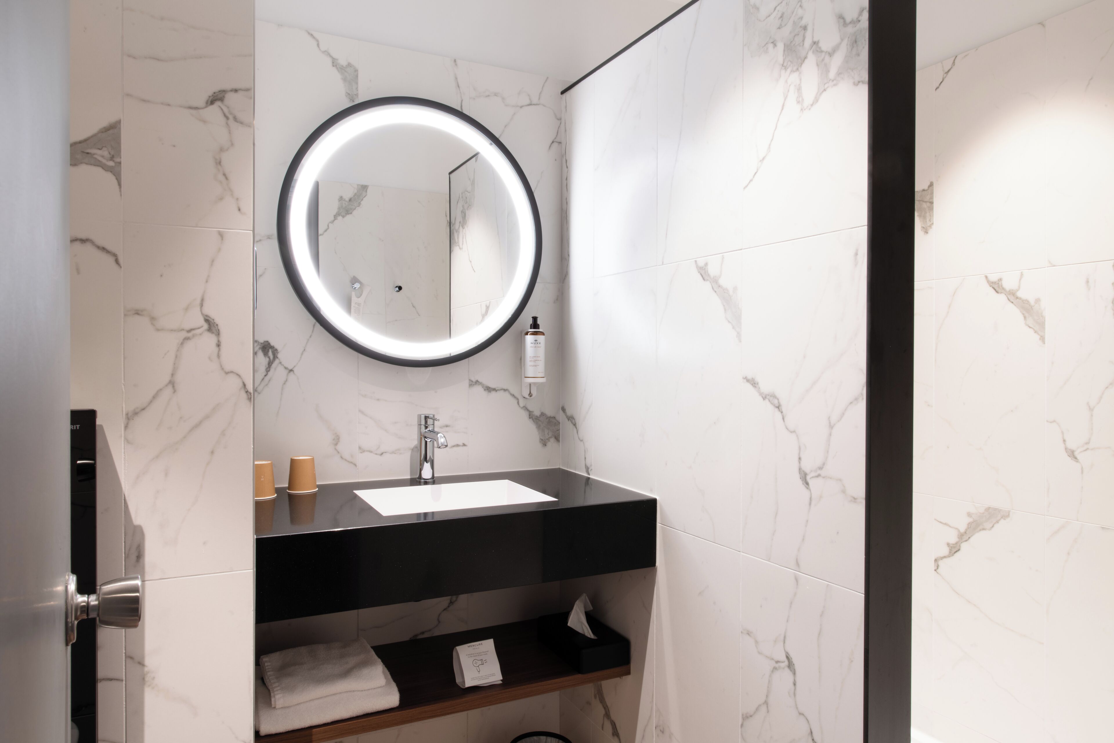 A modern bathroom with a black sink, marble walls, a round mirror with a light, and a towel rack.